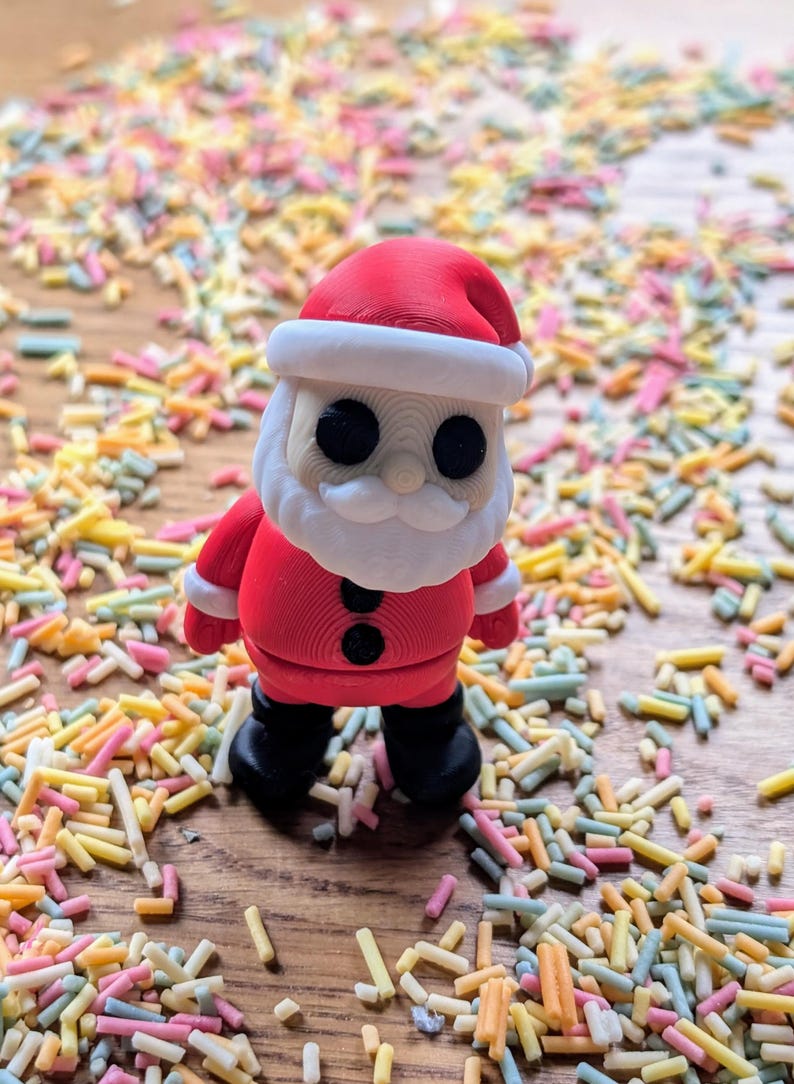 Op de afbeelding: Een rode en witte kerstmanfiguur met een zwarte knoop op zijn jas, staand op een oppervlak bedekt met kleurrijke sprinkels.