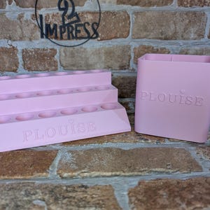 May include: Pink tiered makeup organizer with multiple circular holes and the word "PLouise" embossed on the front. A matching pink square container with the same embossed text is also present. The background is a brick wall.