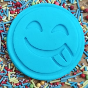 May include: A round, light blue cookie cutter with a smiling face and tongue sticking out. The cutter is surrounded by colorful sprinkles in various shapes and sizes, including blue, red, yellow, and green, on a wooden surface.