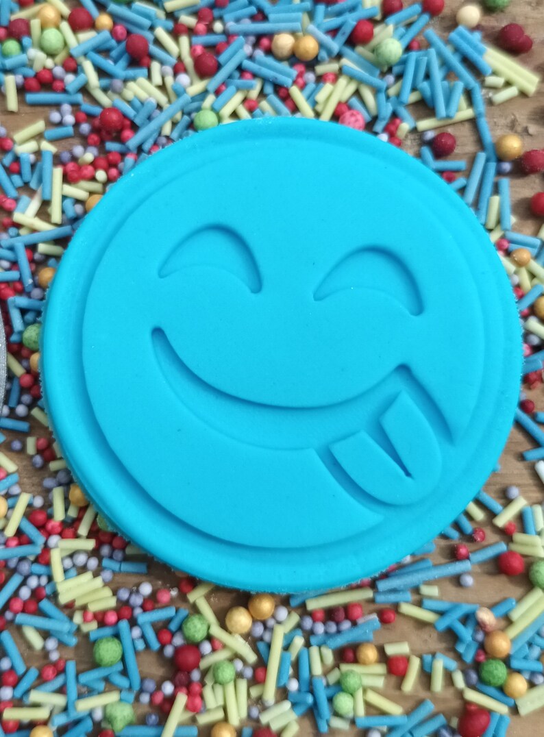 May include: A round, light blue cookie cutter with a smiling face and tongue sticking out. The cutter is surrounded by colorful sprinkles in various shapes and sizes, including blue, red, yellow, and green, on a wooden surface.