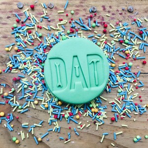 Pu&ograve; includere: Un biscotto rotondo verde chiaro con la scritta "DAD" in rilievo, con disegni a tema utensili all'interno delle lettere. Il biscotto &egrave; circondato da zuccherini colorati su una superficie di legno, creando una scena festosa.