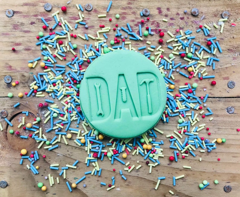 Pu&ograve; includere: Un biscotto rotondo verde chiaro con la scritta "DAD" in rilievo, con disegni a tema utensili all'interno delle lettere. Il biscotto &egrave; circondato da zuccherini colorati su una superficie di legno, creando una scena festosa.