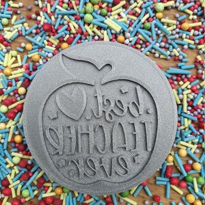 May include: A grey, circular cookie cutter featuring an apple design and the words "Best Teacher Ever". The cutter is surrounded by colourful sprinkles on a wooden surface.
