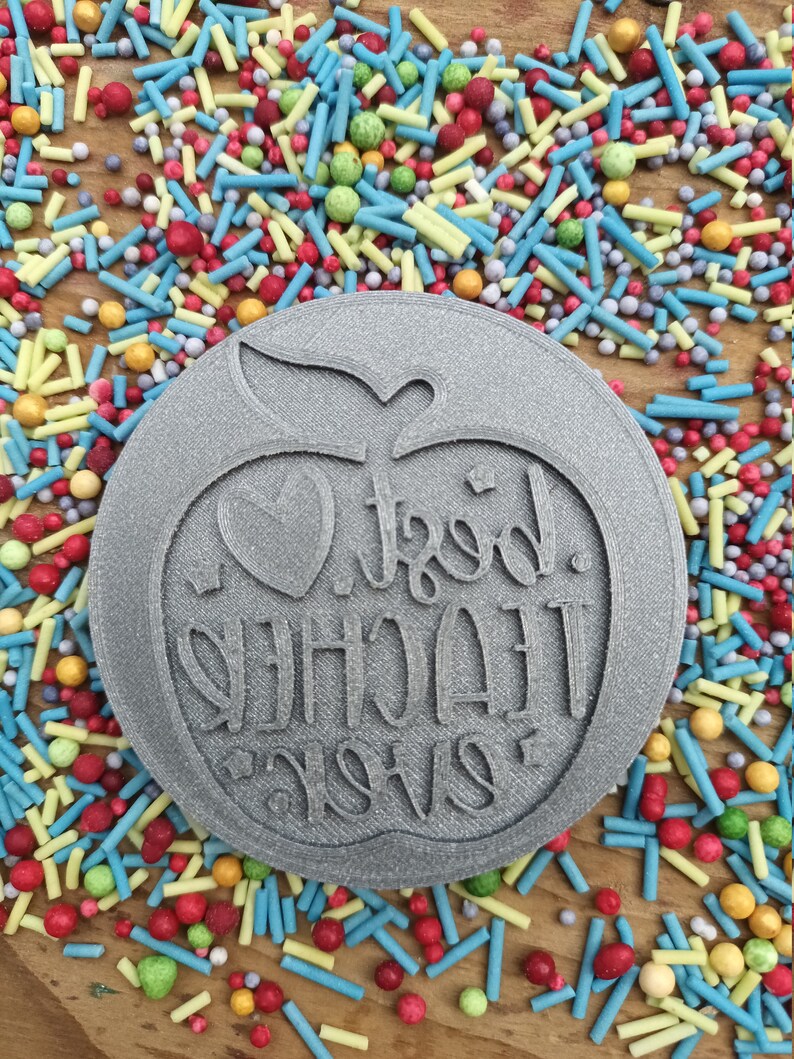 May include: A grey, circular cookie cutter featuring an apple design and the words "Best Teacher Ever". The cutter is surrounded by colourful sprinkles on a wooden surface.
