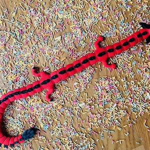 May include: A red and black dragon-shaped toy with a segmented body. The toy is lying on a surface covered in colorful sprinkles.
