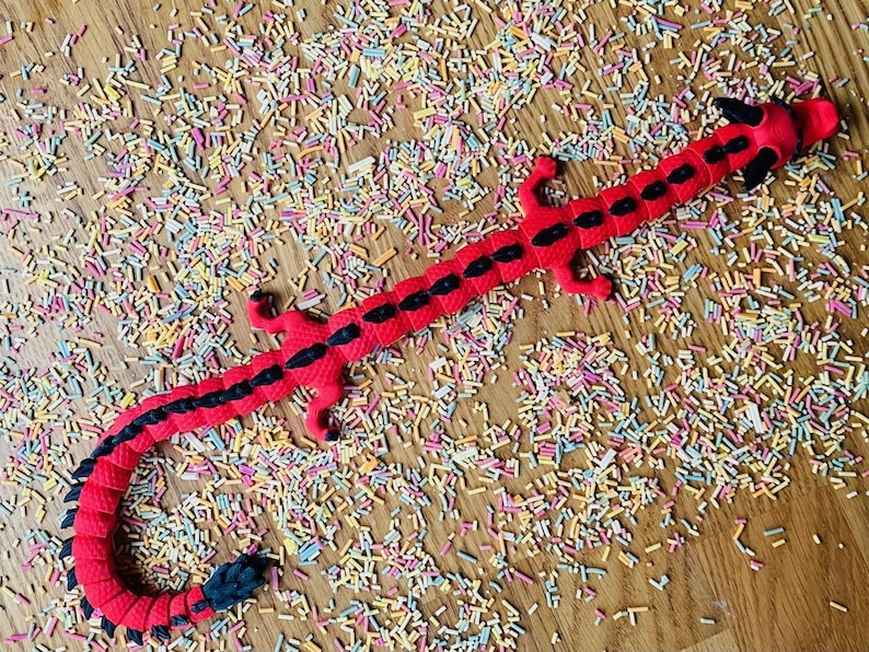 May include: A red and black dragon-shaped toy with a segmented body. The toy is lying on a surface covered in colorful sprinkles.