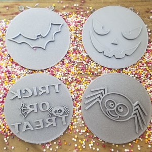 Puede incluir: Cuatro sellos grises para galletas con tem&aacute;tica de Halloween. Los dise&ntilde;os incluyen un murci&eacute;lago, una cara de calabaza, una ara&ntilde;a con las palabras "Trick or Treat" y una ara&ntilde;a sonriente. Los sellos son redondos y est&aacute;n sobre una cama de chispas coloridas.