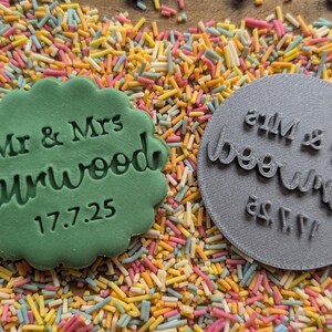 Puede incluir: Una galleta verde con bordes festoneados con el texto "Mr & Mrs Curwood 17.7.25" junto a un sello circular gris con el mismo texto invertido. Los art&iacute;culos descansan sobre un lecho de fideos de colores.