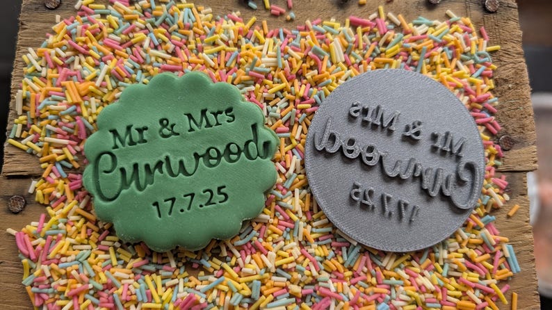Puede incluir: Una galleta verde con bordes festoneados con el texto "Mr & Mrs Curwood 17.7.25" junto a un sello circular gris con el mismo texto invertido. Los art&iacute;culos descansan sobre un lecho de fideos de colores.