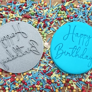 May include: Two round silicone cake stamps with the text "Happy Birthday" in a cursive font. The stamp on the left is gray and the stamp on the right is blue.