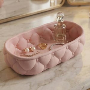 May include: A pink, oval quilted tray displaying jewellery and a perfume bottle. The tray features a button-tufted design. The jewellery includes pearl earrings, a gold ring, and a bracelet. The perfume bottle is clear with a decorative stopper.