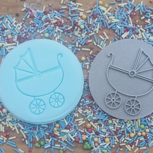 May include: Two round cookie cutters, one light blue and one gray, each featuring a baby carriage design. The cutters are surrounded by colorful sprinkles. The baby carriage design includes a hood and four wheels.