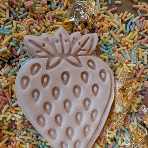 May include: A light pink strawberry-shaped confection with detailed seed impressions, resting on a bed of colorful sprinkles. Gold leaf accents add a touch of elegance to this dessert-themed image.