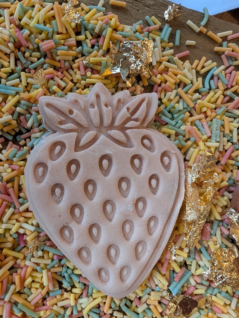 May include: A light pink strawberry-shaped confection with detailed seed impressions, resting on a bed of colorful sprinkles. Gold leaf accents add a touch of elegance to this dessert-themed image.