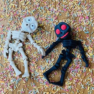 May include: Two skeleton toys, one white and one black, are lying on a wooden surface covered in colorful sprinkles. The white skeleton has large, round eyes, while the black skeleton has red eyes.