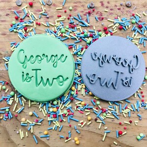 Op de afbeelding: Twee ronde koekjesvormen, een groene en een grijze, met de tekst "George is Two" op de groene vorm en "George is Two" op de grijze vorm. De vormen zijn omringd door kleurrijke sprinkles.