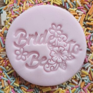 May include: A light pink, circular mold with the words "Bride To Be" and floral designs. The mold is surrounded by colorful sprinkles, creating a festive and celebratory theme. Ideal for cake decorating.