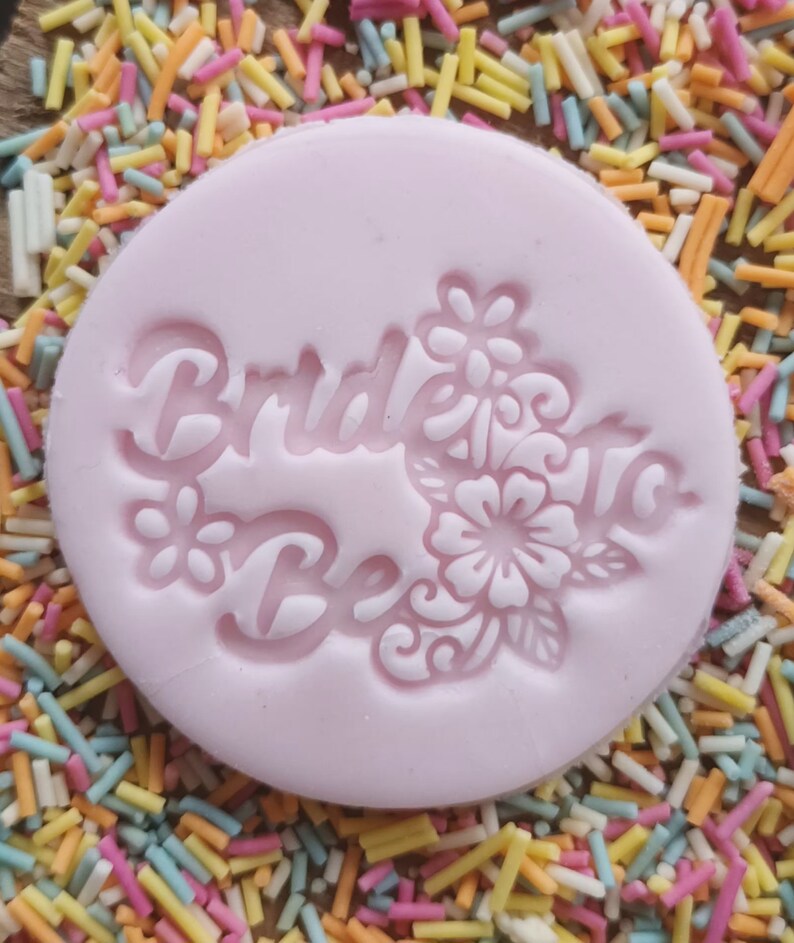 May include: A light pink, circular mold with the words "Bride To Be" and floral designs. The mold is surrounded by colorful sprinkles, creating a festive and celebratory theme. Ideal for cake decorating.