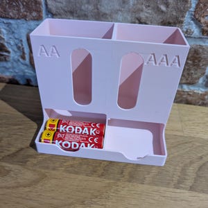 May include: A light pink battery organizer with compartments for AA and AAA batteries. The organizer has a tray at the bottom and holds two Kodak AA batteries. The text "AA" and "AAA" are visible on the organizer.