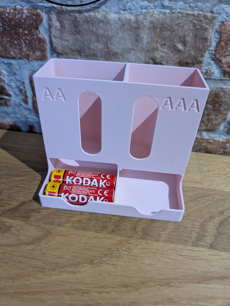 May include: A light pink battery organizer with compartments for AA and AAA batteries. The organizer has a tray at the bottom and holds two Kodak AA batteries. The text "AA" and "AAA" are visible on the organizer.