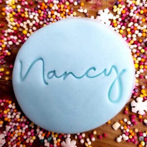 May include: A light blue round cake topper with the name "Nancy" written in cursive script. The topper is surrounded by colorful sprinkles.