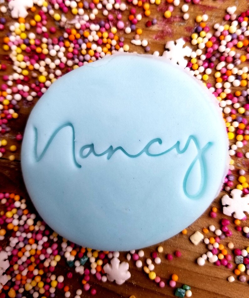 May include: A light blue round cake topper with the name "Nancy" written in cursive script. The topper is surrounded by colorful sprinkles.