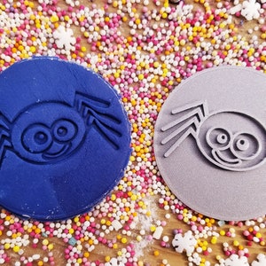 Puede incluir: Dos sellos circulares para galletas con un dise&ntilde;o de ara&ntilde;a de dibujos animados. Un sello es azul marino, el otro gris. Los sellos est&aacute;n rodeados de chispas de colores sobre una superficie de madera, creando una escena l&uacute;dica y festiva.
