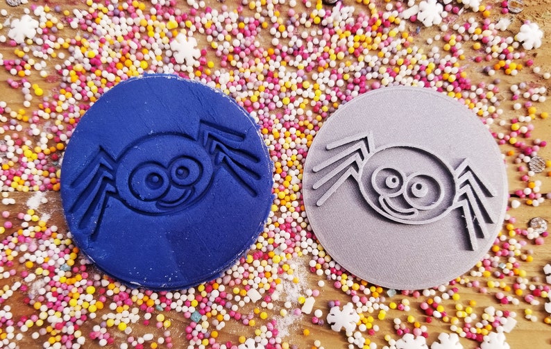 Puede incluir: Dos sellos circulares para galletas con un dise&ntilde;o de ara&ntilde;a de dibujos animados. Un sello es azul marino, el otro gris. Los sellos est&aacute;n rodeados de chispas de colores sobre una superficie de madera, creando una escena l&uacute;dica y festiva.
