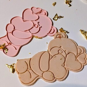 May include: Two pink and beige bear-shaped cookie cutters with heart details. Gold leaf flakes are scattered around the cutters. The cutters are made of a flexible material, designed for baking or crafting.