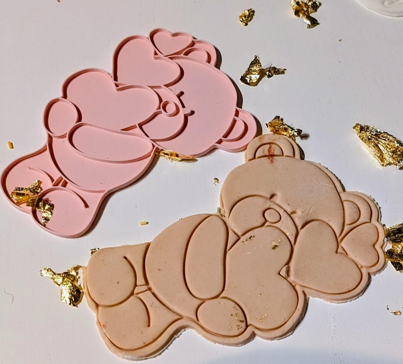 May include: Two pink and beige bear-shaped cookie cutters with heart details. Gold leaf flakes are scattered around the cutters. The cutters are made of a flexible material, designed for baking or crafting.