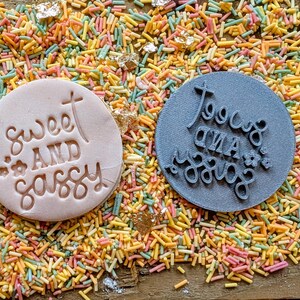 May include: Two round cookie stamps, one light pink and one gray, with the words "Sweet and Sassy" and "Too Cute and Sassy" embossed on them. The stamps are surrounded by colorful sprinkles and gold flakes.