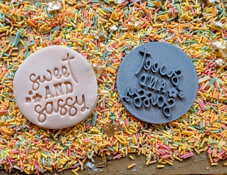 May include: Two round cookie stamps, one light pink and one gray, with the words "Sweet and Sassy" and "Too Cute and Sassy" embossed on them. The stamps are surrounded by colorful sprinkles and gold flakes.