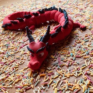 May include: A red plastic dragon toy with black accents and googly eyes, lying on a surface covered in colorful sprinkles.