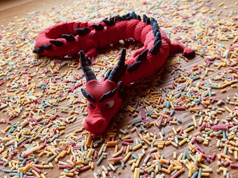 May include: A red plastic dragon toy with black accents and googly eyes, lying on a surface covered in colorful sprinkles.