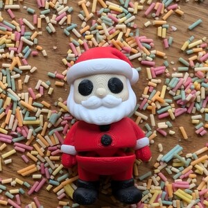 Op de afbeelding: Een rode en witte kerstmanfiguur met een zwarte riem en zwarte laarzen. Het figuur is omringd door kleurrijke sprinkels.
