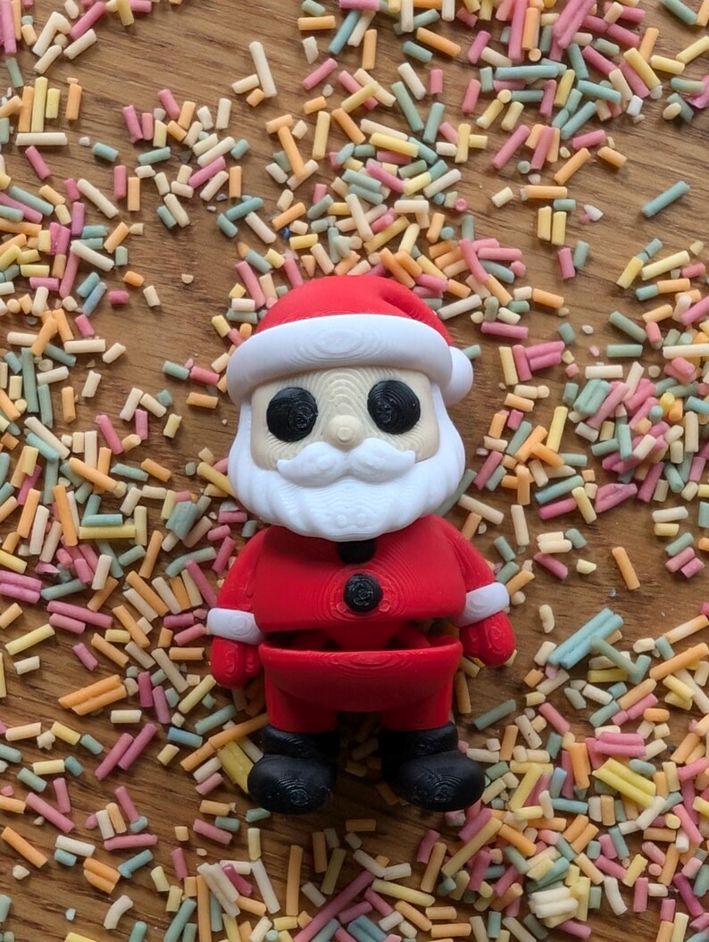 Op de afbeelding: Een rode en witte kerstmanfiguur met een zwarte riem en zwarte laarzen. Het figuur is omringd door kleurrijke sprinkels.
