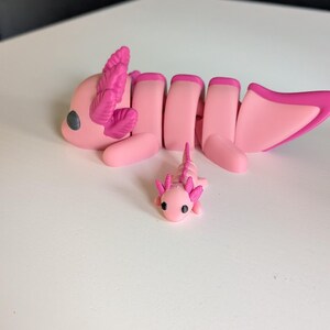 May include: Two pink axolotl figurines, one larger than the other, with detailed features and pink gills. The larger axolotl is lying down with its tail curled up, while the smaller axolotl is standing upright.