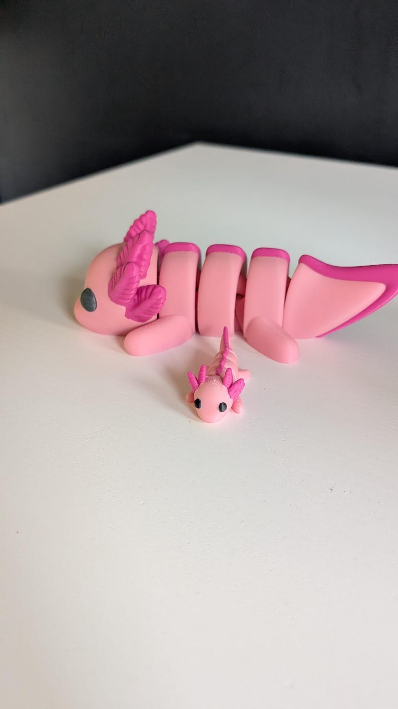 May include: Two pink axolotl figurines, one larger than the other, with detailed features and pink gills. The larger axolotl is lying down with its tail curled up, while the smaller axolotl is standing upright.