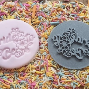 May include: Two round molds for cake decorating. One is pink and embossed with the words "Bride to Be" and floral designs. The other is gray and has a floral design with the words "Bride to Be" cut out. The molds are surrounded by colorful sprinkles.