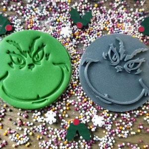 May include: Two green and gray Grinch cookie cutters on a wooden surface with colorful sprinkles. The Grinch's face is visible on both cutters.