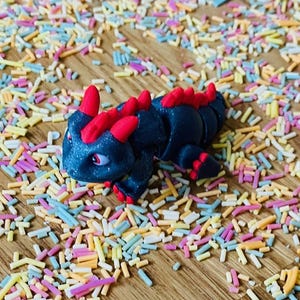May include: A small, black, segmented toy dragon with red spines and a glittery finish. The dragon is posed on a wooden surface covered in colorful sprinkles.
