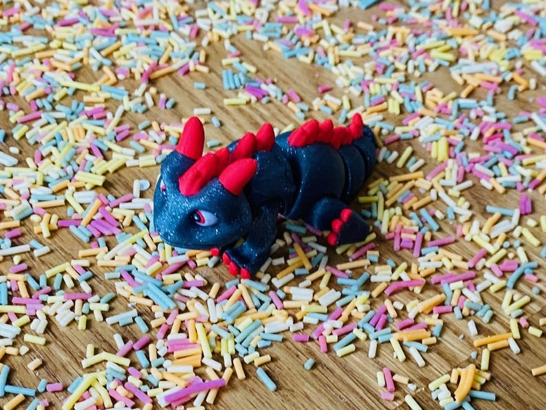 May include: A small, black, segmented toy dragon with red spines and a glittery finish. The dragon is posed on a wooden surface covered in colorful sprinkles.