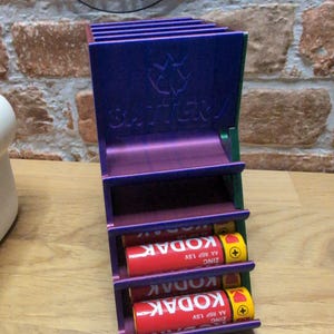 May include: A multi-colored battery holder with the word "BATTERY" and a graphic. The holder contains three red and yellow Kodak AA batteries. The holder is made of purple, green, and blue metal.