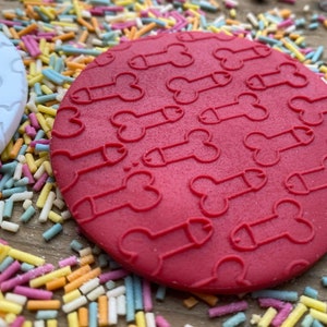 May include: A red, circular coaster with a repeating pattern of phallus shapes. A white coaster with a similar design is visible to the left. The coasters are surrounded by colorful sprinkles, suggesting a playful or humorous theme.