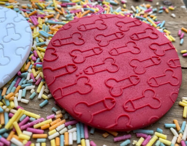 May include: A red, circular coaster with a repeating pattern of phallus shapes. A white coaster with a similar design is visible to the left. The coasters are surrounded by colorful sprinkles, suggesting a playful or humorous theme.