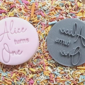 Puede incluir: Dos sellos circulares para pasteles, uno rosa y otro gris, con las palabras "Alice turns One" en fuente cursiva. Los sellos est&aacute;n rodeados de chispas de colores, lo que sugiere un tema de celebraci&oacute;n. Ideal para un primer cumplea&ntilde;os.