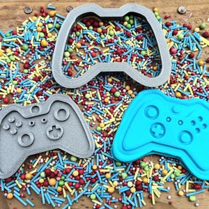 以下が含まれることがあります： カラフルなスプリンクルを背景に、グレーのプラスチック製のゲームコントローラーのクッキーカッター2つと、青いシリコン製のゲームコントローラーのモールド1つ