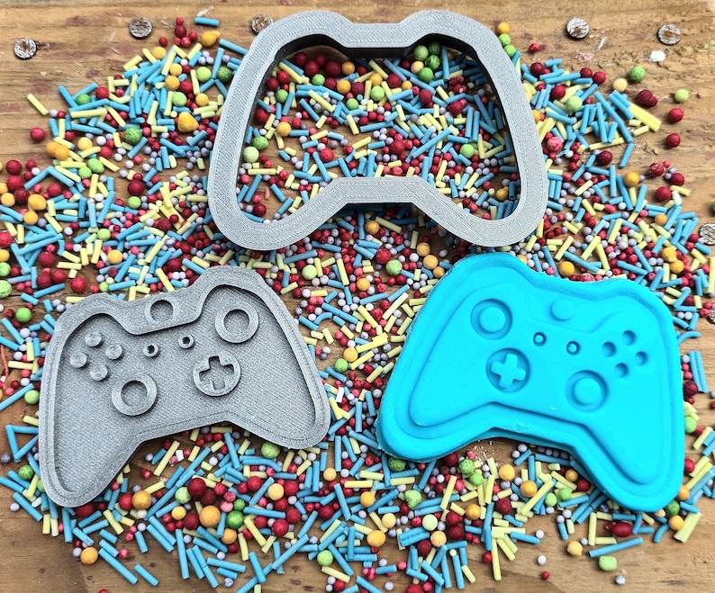 以下が含まれることがあります： カラフルなスプリンクルを背景に、グレーのプラスチック製のゲームコントローラーのクッキーカッター2つと、青いシリコン製のゲームコントローラーのモールド1つ