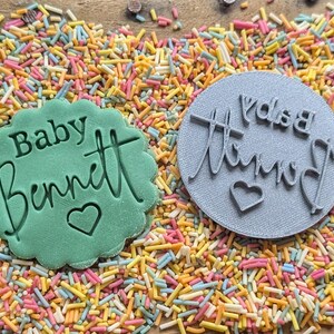 K&ouml;nnte beinhalten: Zwei Kekssiegel mit Text und Herzdetails auf einem Bett aus bunten Streuseln. Ein Stempel ist gr&uuml;n und tr&auml;gt die Aufschrift "Baby Bennett" mit einem Herzen. Der andere ist grau und tr&auml;gt die Aufschrift "Bennett's Baby" mit einem Herzen.