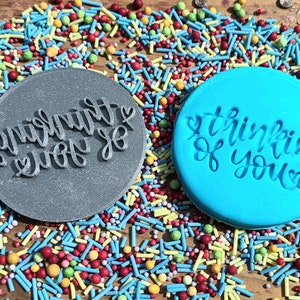 May include: Two round cookie stamps, one gray and one blue, with the phrases "thinking of you" and "everything ever so" in cursive. The stamps are surrounded by colorful sprinkles, perfect for baking and crafting.